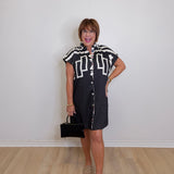 Woman wearing a black and white patterned dress holding a black handbag against a plain white wall.