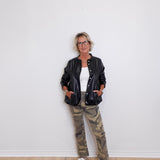 Person wearing a black leather jacket, white shirt, and camouflage pants against a white wall.