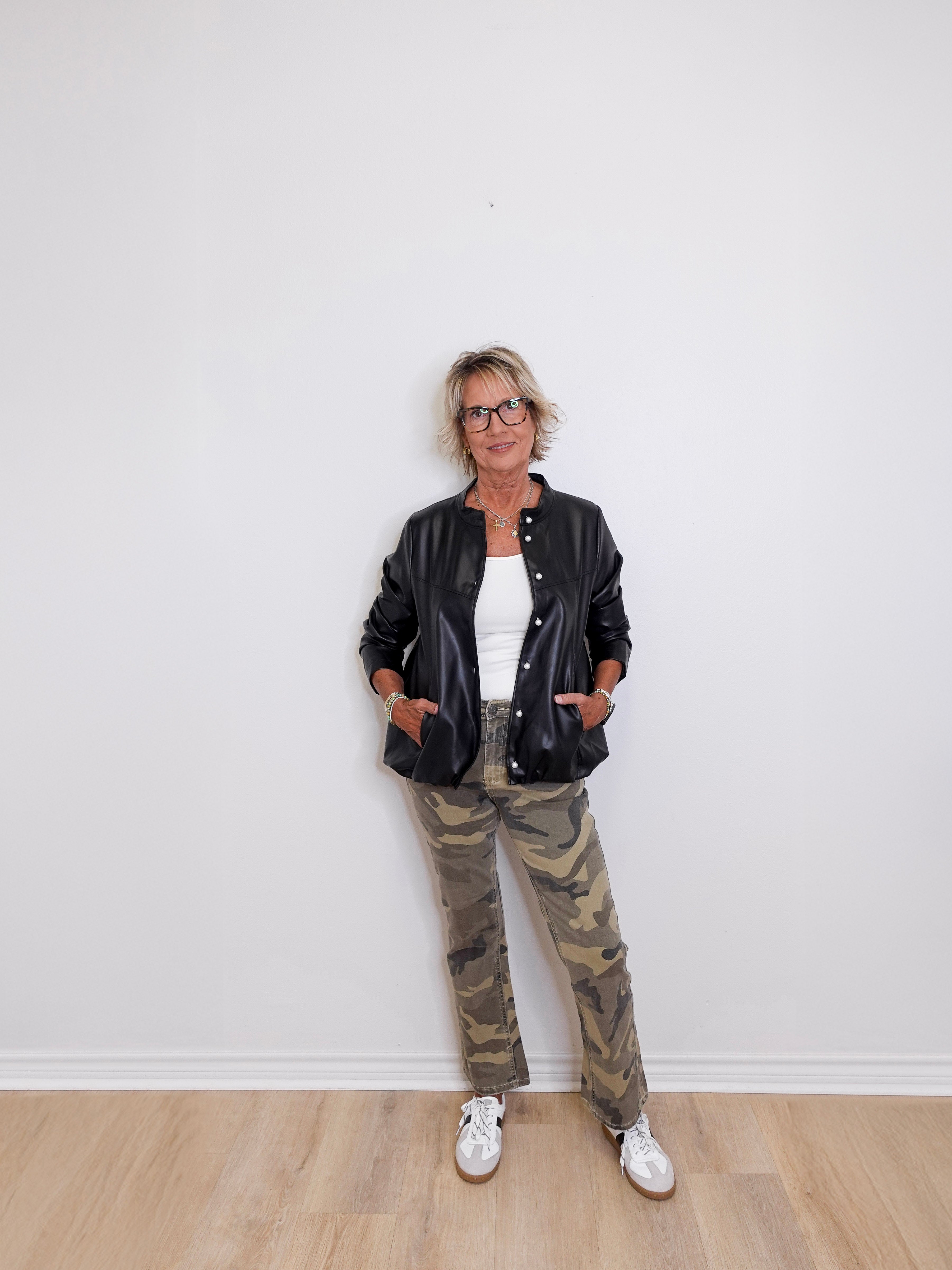 Person wearing a black leather jacket, white shirt, and camouflage pants against a white wall.