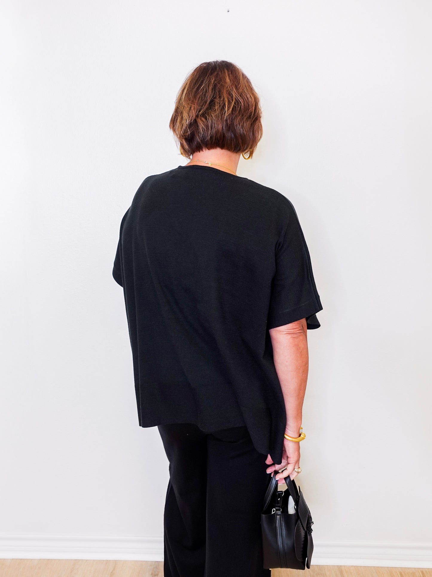 Person wearing a black outfit holding a black handbag against a white wall