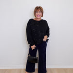 Woman wearing a black sweater and wide-leg pants against a white wall.