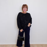 Woman wearing a black sweater and wide-leg pants against a white wall.