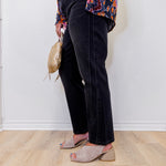 Person wearing a colorful patterned top, black pants, and beige sandals against a white wall.