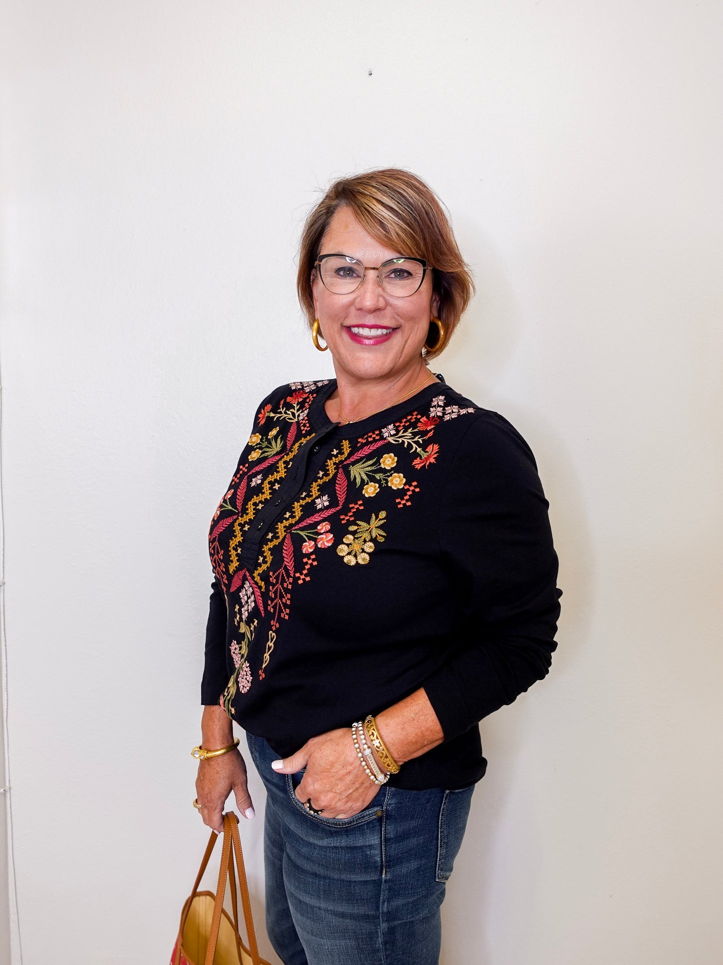 Woman wearing a black sweater with floral embroidery, holding a handbag against a white wall.