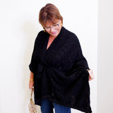 Woman wearing a black poncho and blue jeans against a white wall.