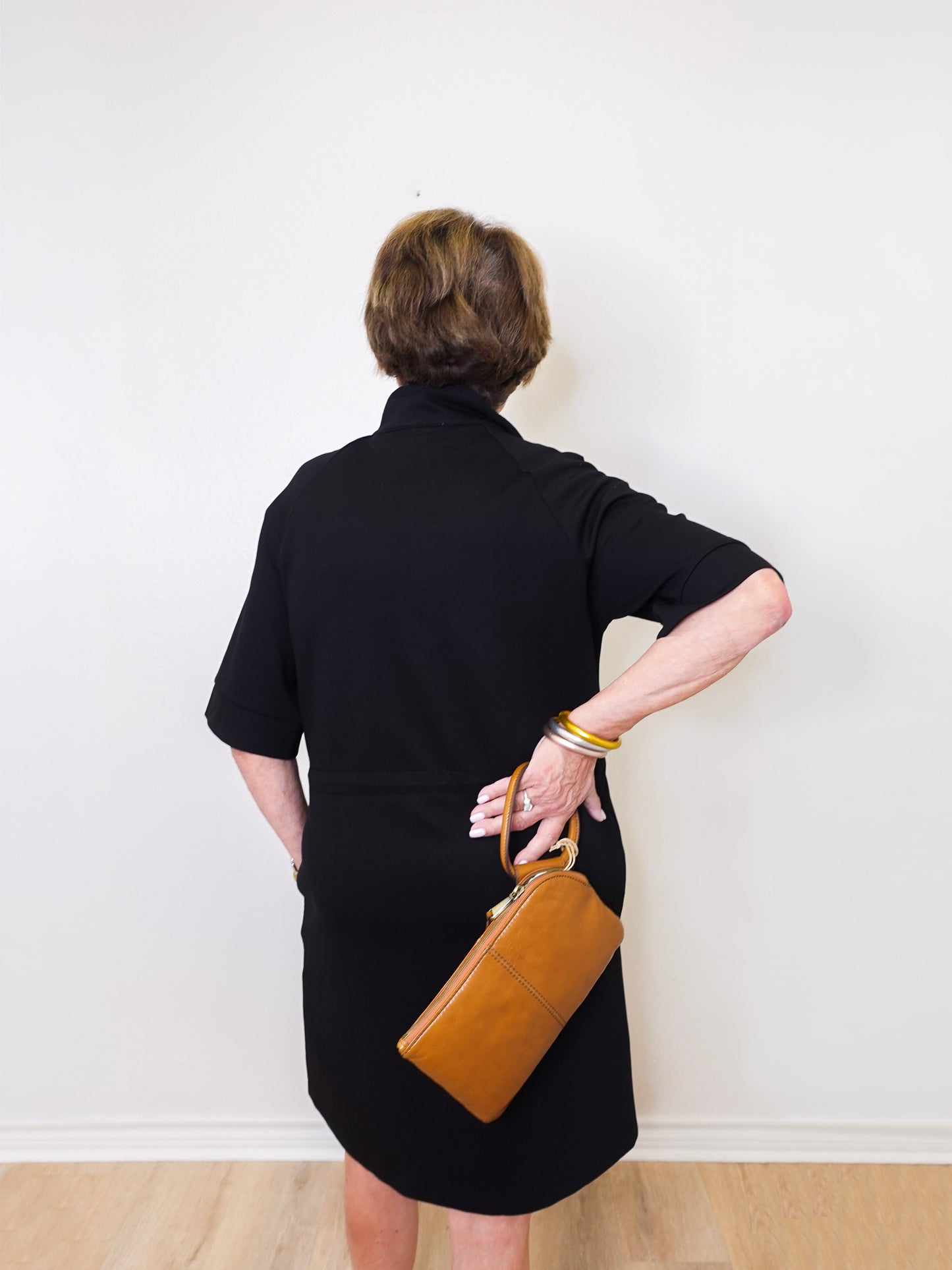 Person wearing a black dress holding a brown leather bag against a white wall.