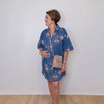Woman wearing a blue floral dress with a beige bag against a white wall.