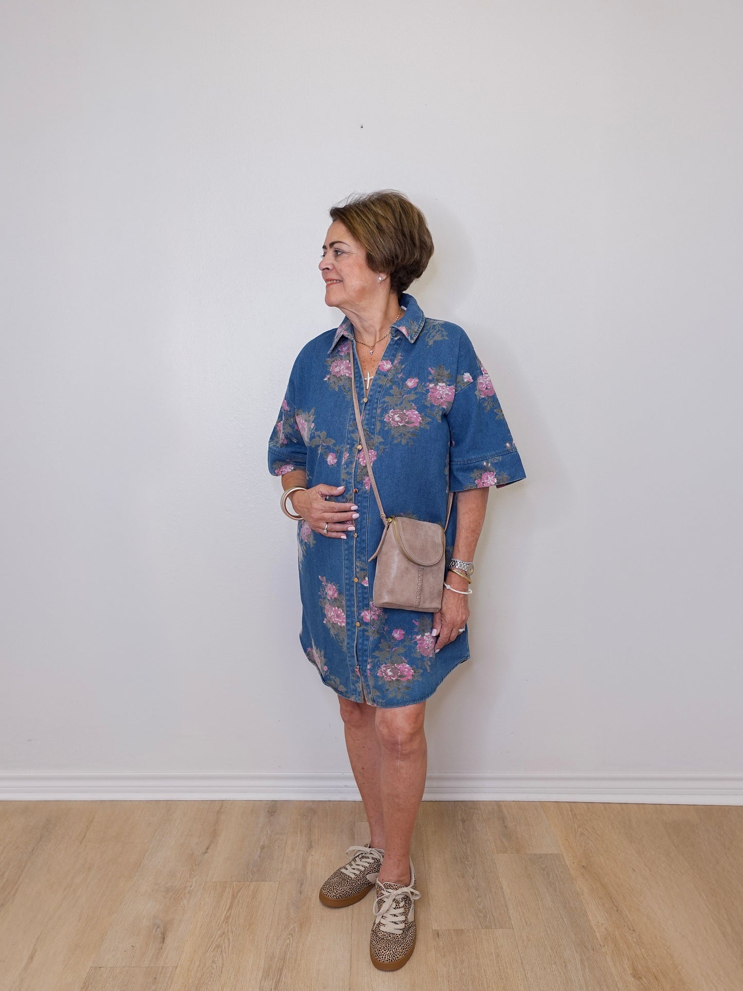 Woman wearing a blue floral dress with a beige bag against a white wall.