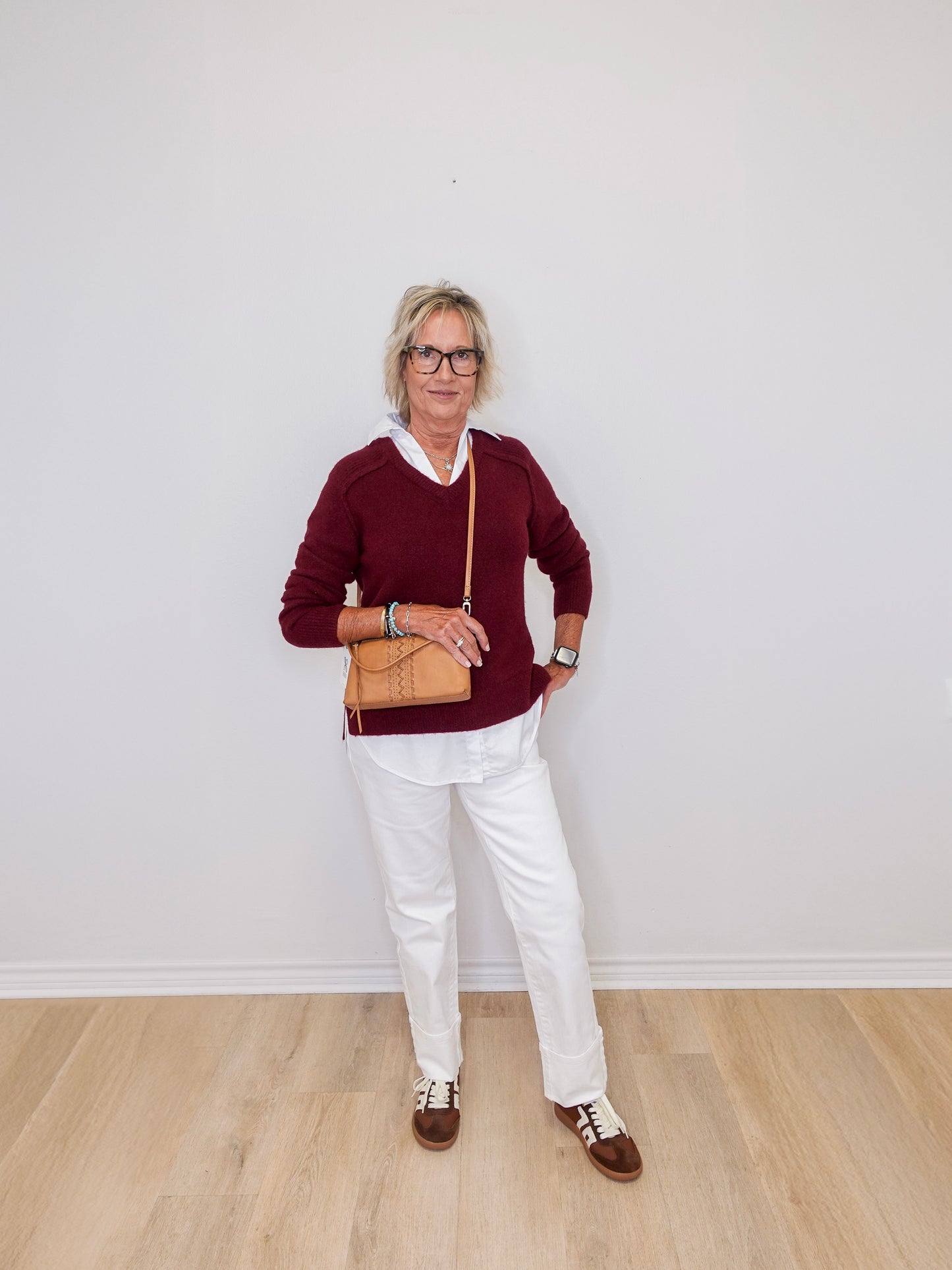 Person wearing a burgundy sweater, white pants, and brown shoes against a plain white wall.