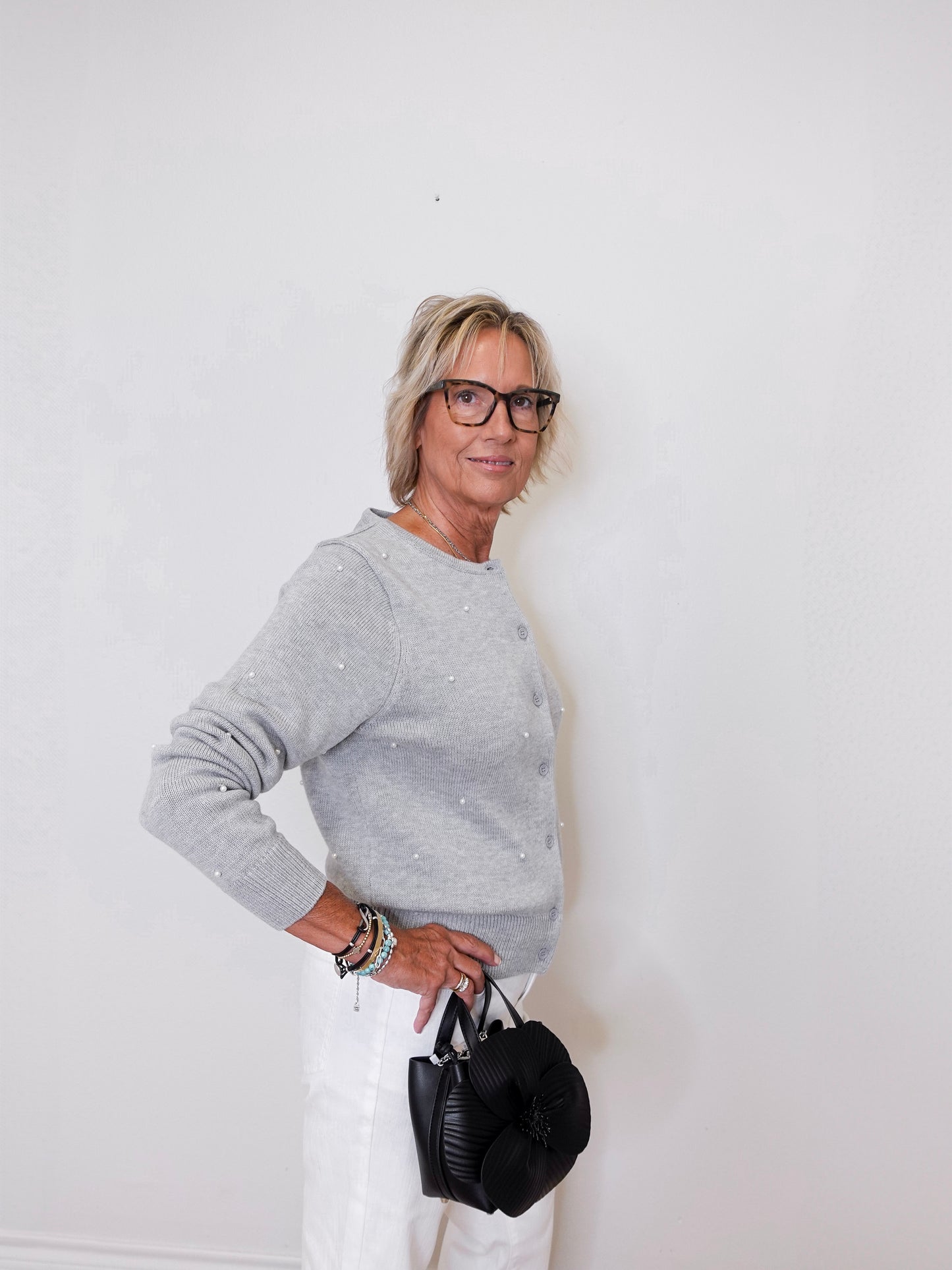 Woman wearing a gray sweater and white pants, holding a black handbag against a white background