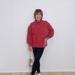 Woman wearing a red sweater with a bow, black pants, and white sneakers against a white wall.