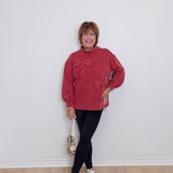Woman wearing a red sweater with a bow, black pants, and white sneakers against a white wall.