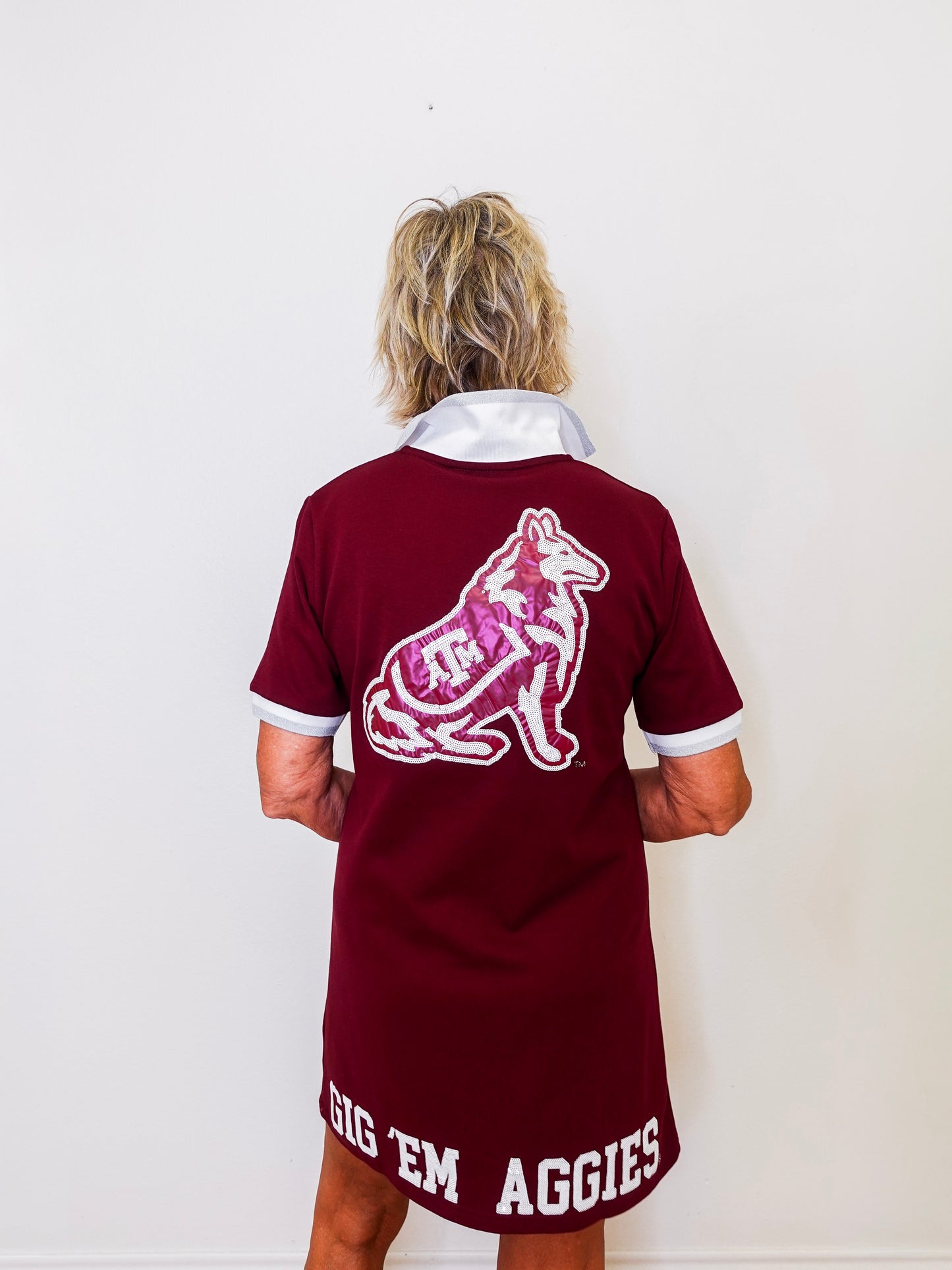 Person wearing a maroon jersey with a logo and text on the back, standing against a white wall.