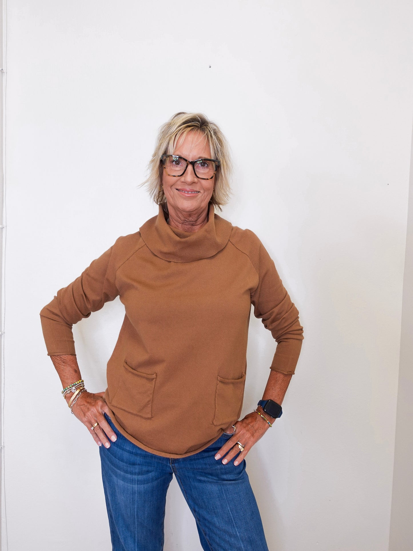 Casual outfit featuring a tan cowl neck sweater and blue jeans on a woman by a white wall.