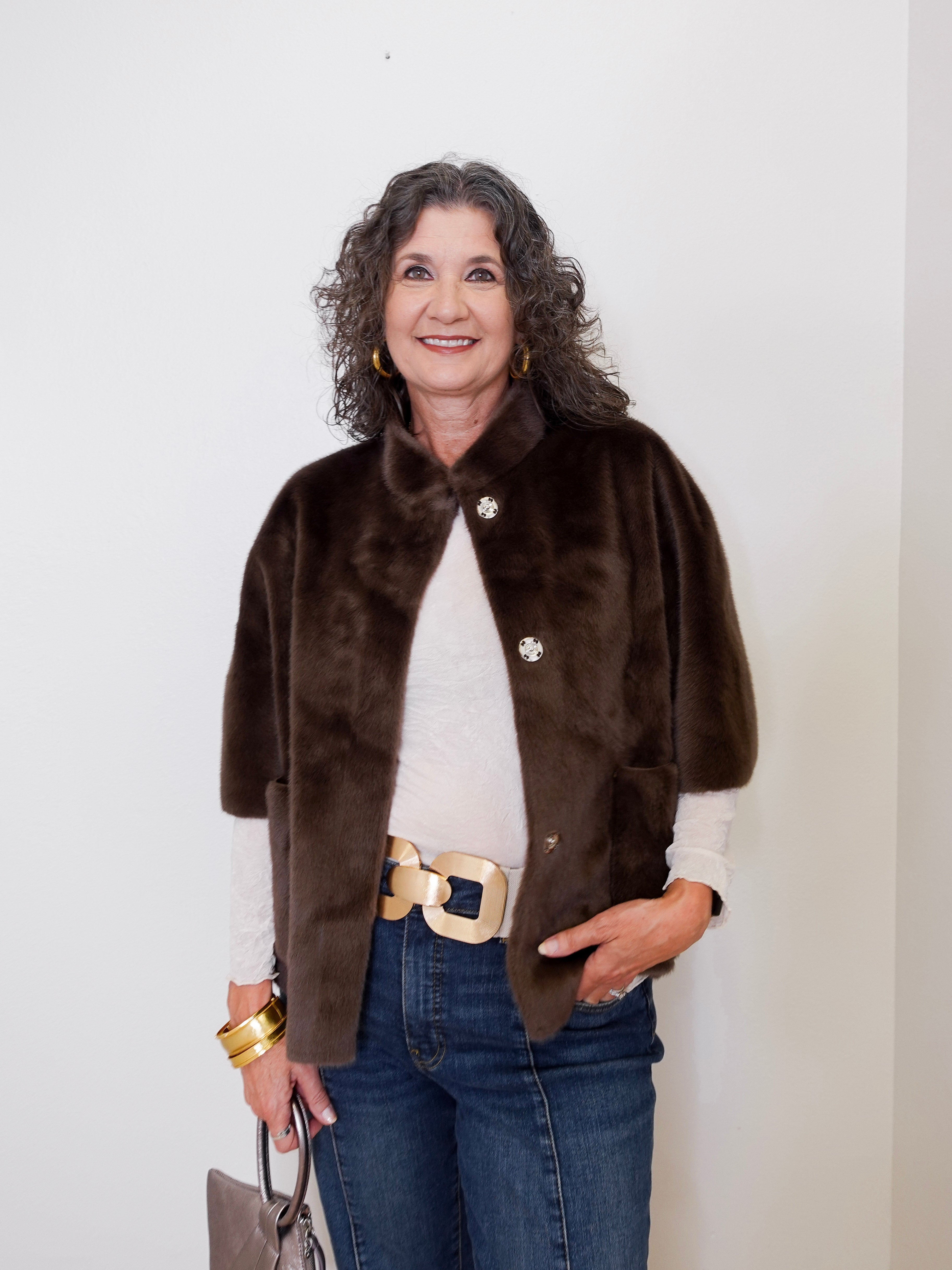 Woman wearing a brown fur vest over a white shirt with blue jeans against a plain background
