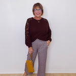 Woman wearing a burgundy sweater with lace sleeves and plaid pants, holding a yellow handbag against a white wall.