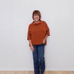 Woman wearing a brown top and blue jeans standing against a white wall.