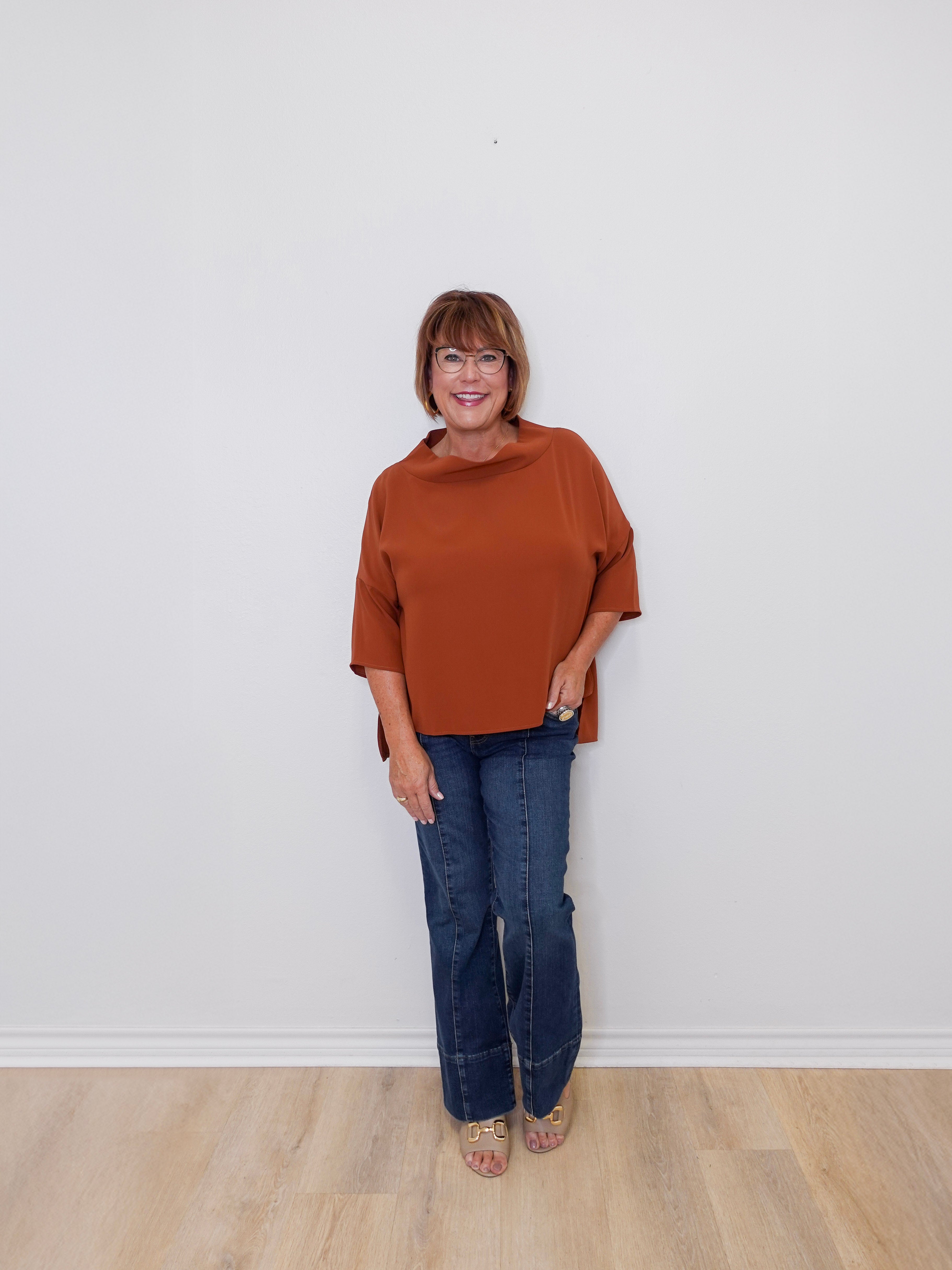 Woman wearing a brown top and blue jeans standing against a white wall.
