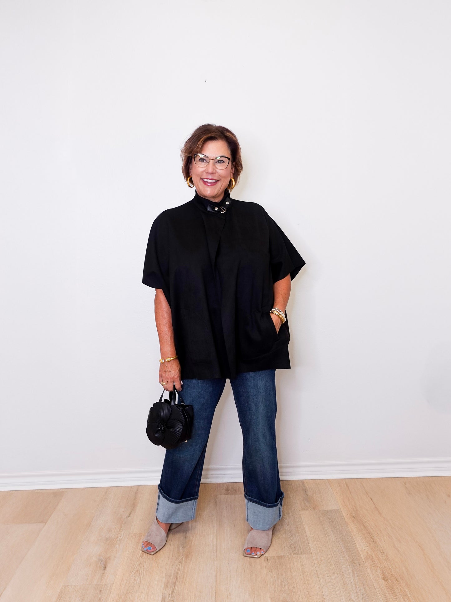 Woman wearing a black top and blue jeans standing against a white wall.