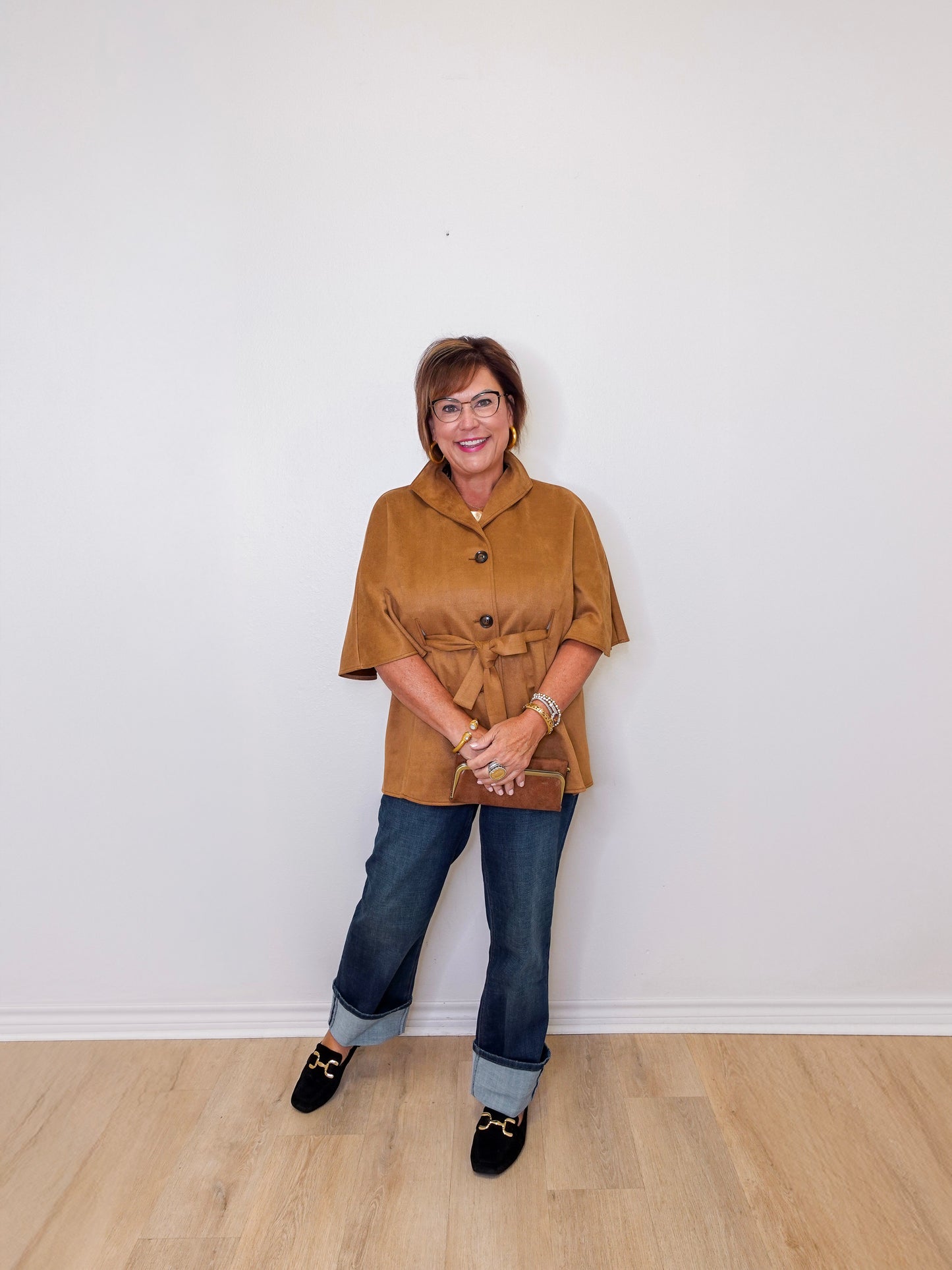 Woman wearing a brown jacket and blue jeans standing against a white wall.