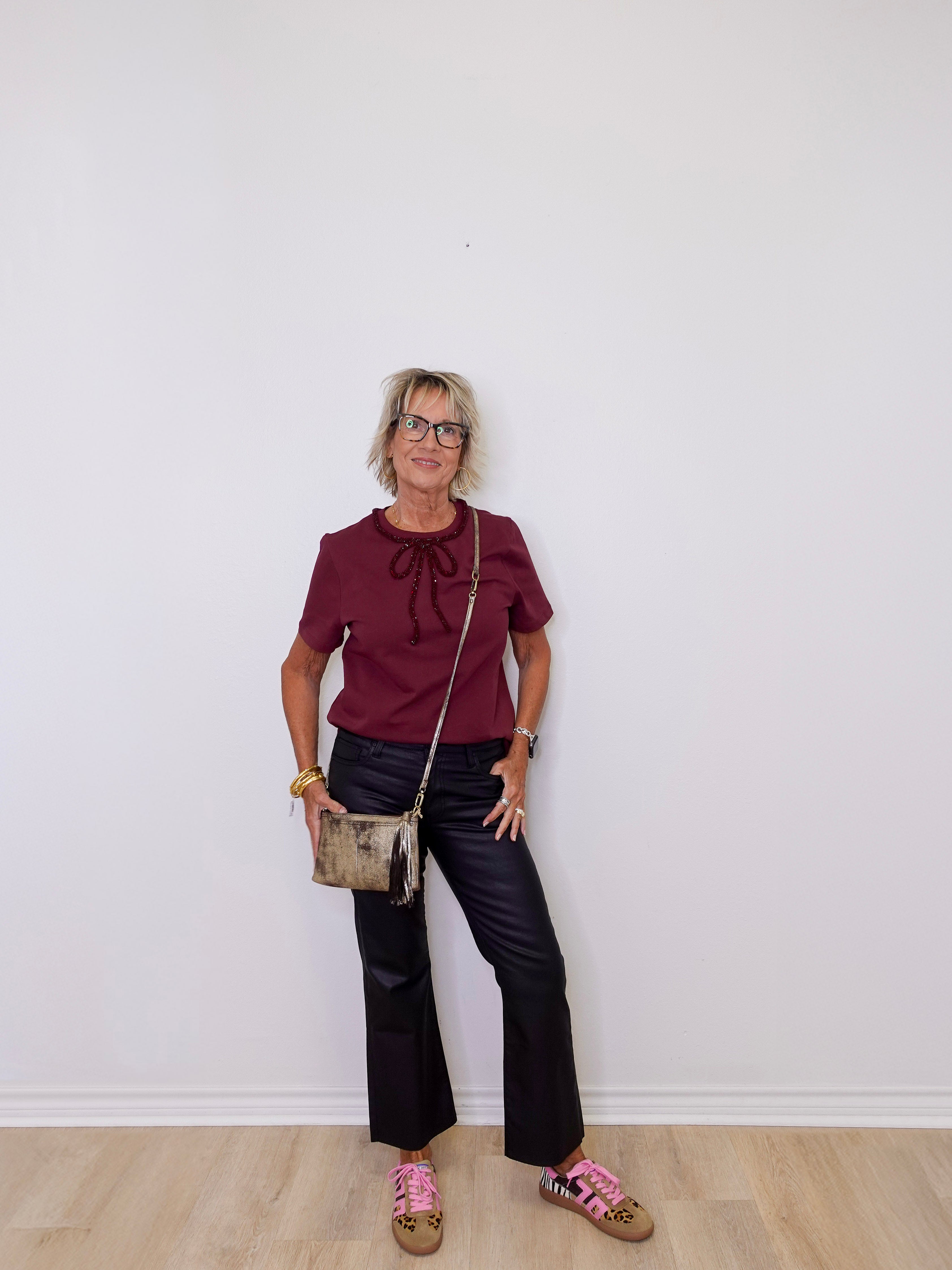 Woman wearing a burgundy top and black pants, standing against a white wall.
