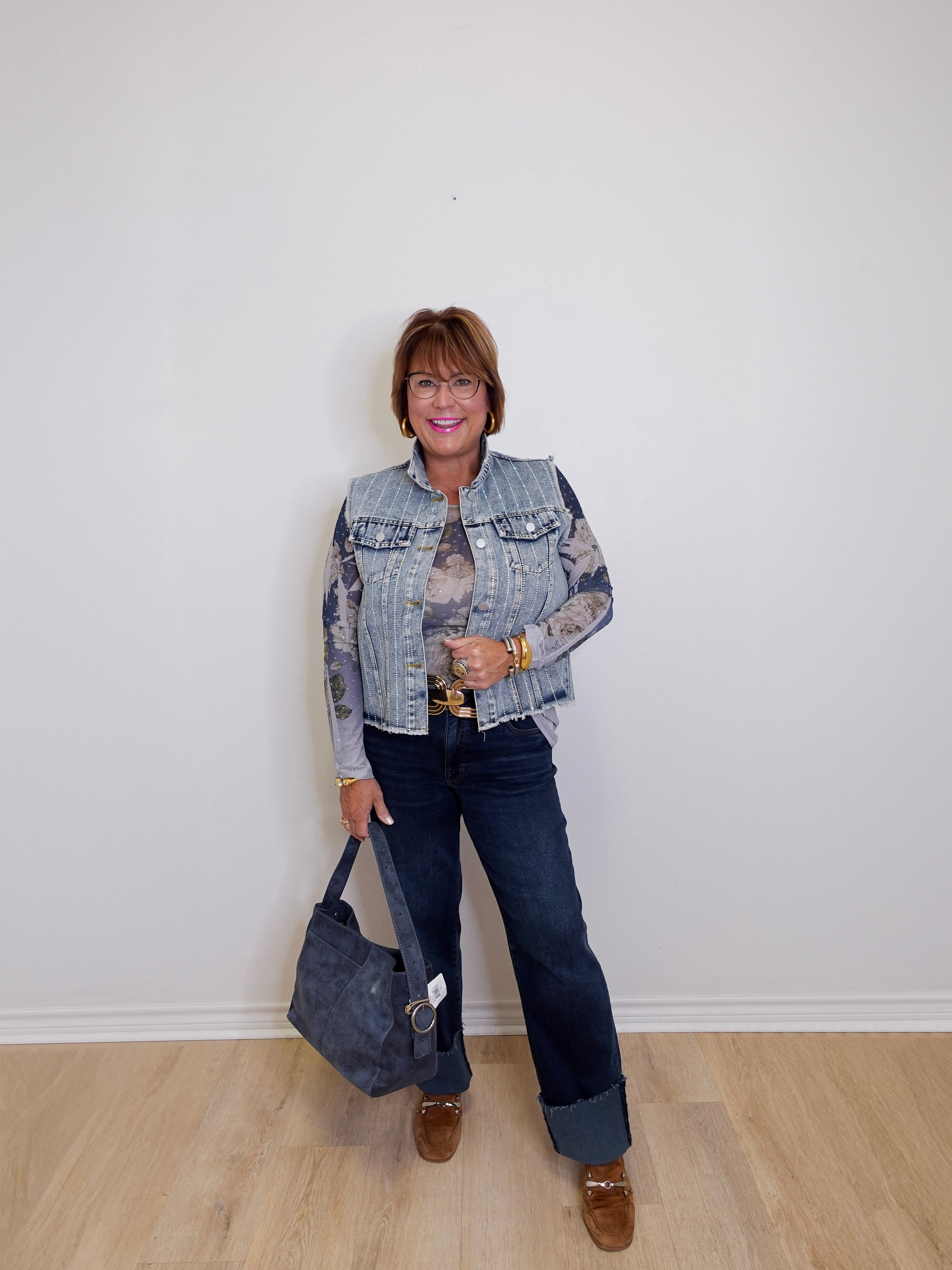 Woman wearing a denim jacket and jeans, holding a gray bag against a white wall.