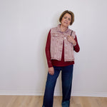 Woman wearing a red shirt, patterned vest, blue jeans, and white sneakers against a white wall.