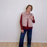 Woman wearing a red shirt, patterned vest, blue jeans, and white sneakers against a white wall.