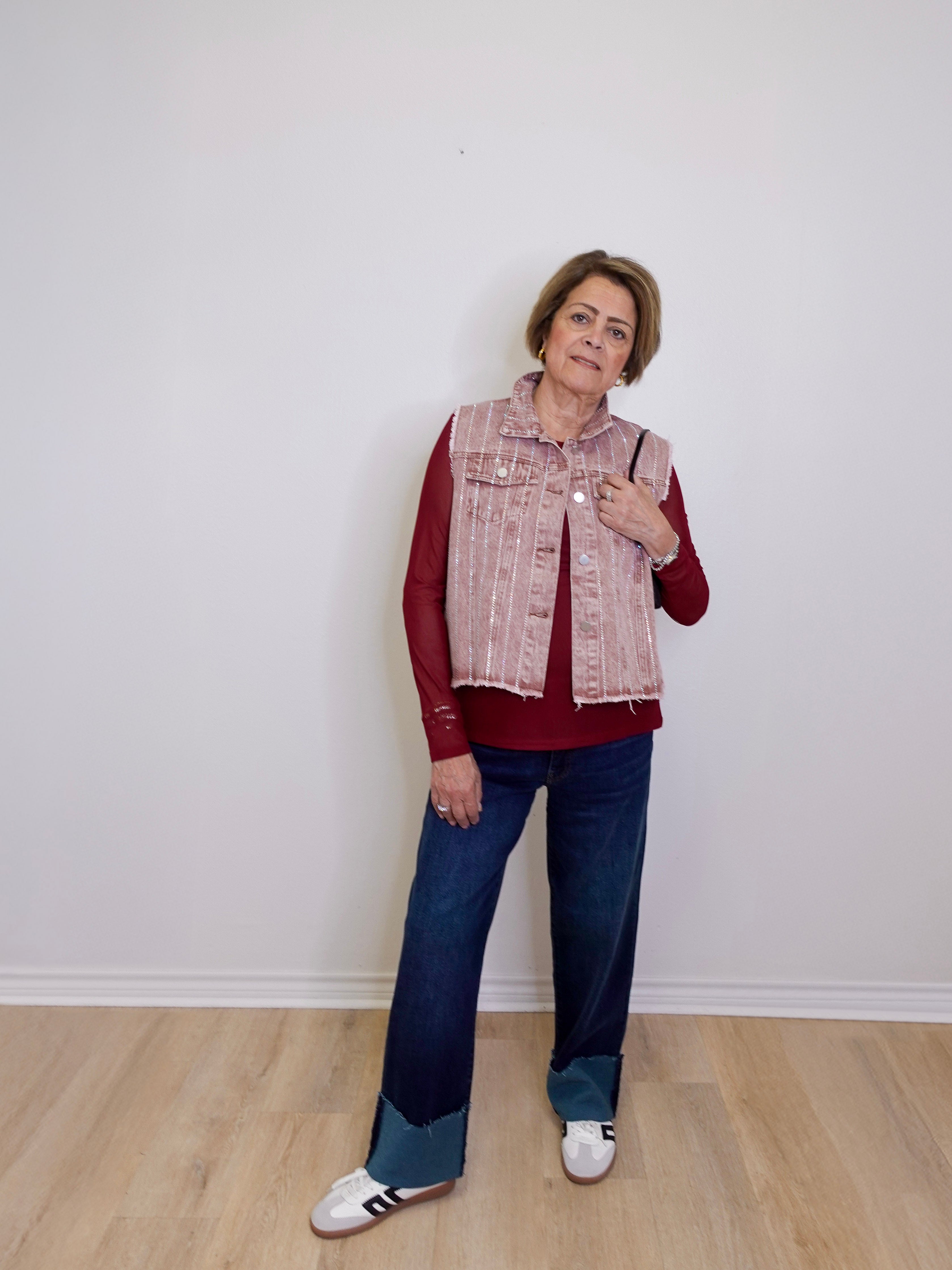 Woman wearing a red shirt, patterned vest, blue jeans, and white sneakers against a white wall.