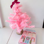 Small pink artificial Christmas tree with a purple bow next to a decorative box on a white surface.