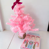 Small pink artificial Christmas tree with a purple bow next to a decorative box on a white surface.