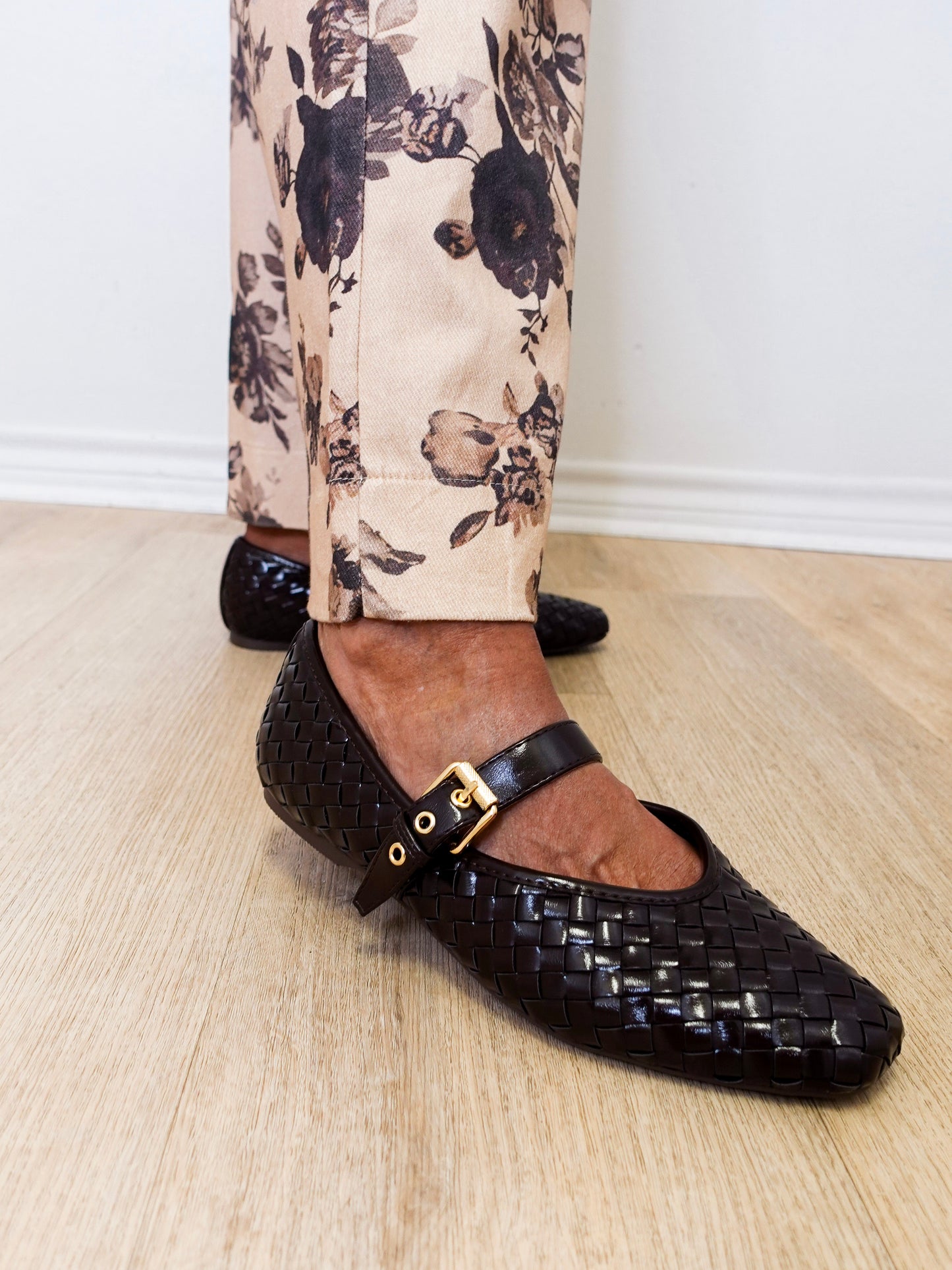 Black woven flats with a gold buckle worn with floral pants on a wooden floor.