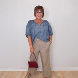 Woman wearing a blue blouse and beige pants, holding a red handbag against a white wall.
