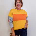 Woman wearing a yellow, orange, and gray sweater with dark jeans against a white background
