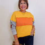 Woman wearing a yellow, orange, and gray sweater with dark jeans against a white background