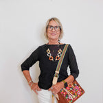 Woman wearing a black top, white pants, and carrying a colorful handbag against a plain background