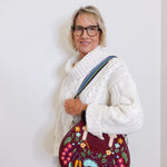 Woman wearing a white knitted sweater holding a colorful embroidered bag against a plain background
