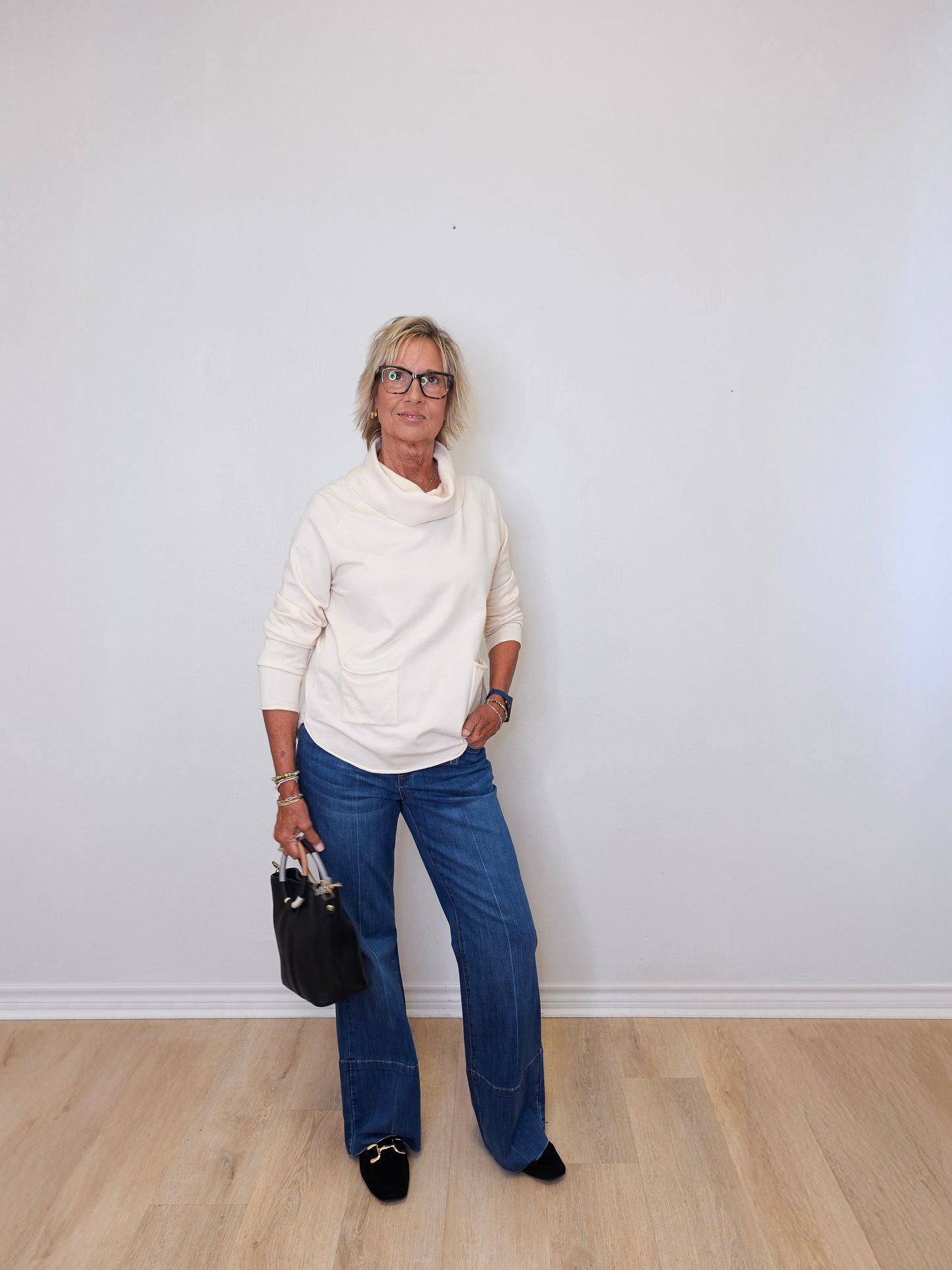 A woman wearing a cowl neck sweater in cream with blue pants against a white wall.