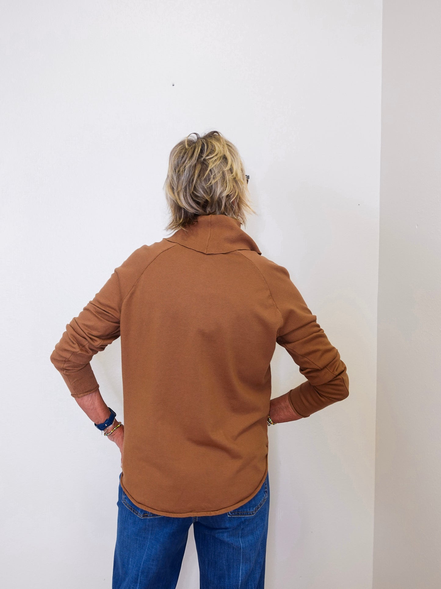 A woman wearing a tan cowl neck sweater with blue jeans against a white wall.