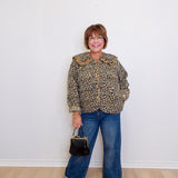 Person wearing a leopard print jacket and blue jeans, holding a black handbag against a white wall.