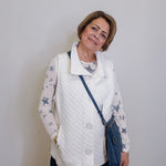 Woman wearing a white quilted vest over a star-patterned shirt with a blue crossbody bag against a plain background.