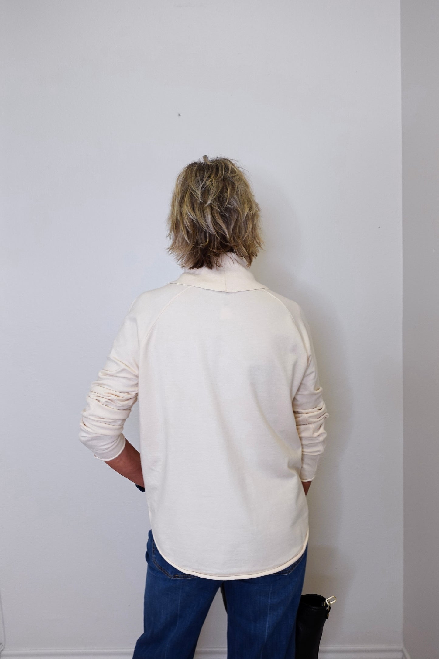 A woman wearing a cowl neck sweater in cream with blue pants against a white wall.