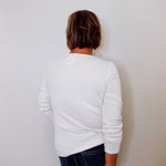 Person wearing a white long sleeve top and dark jeans against a plain wall