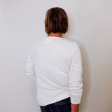 Person wearing a white long sleeve top and dark jeans against a plain wall