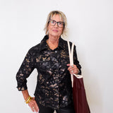 Woman wearing a black patterned shirt and holding a brown bag against a white wall