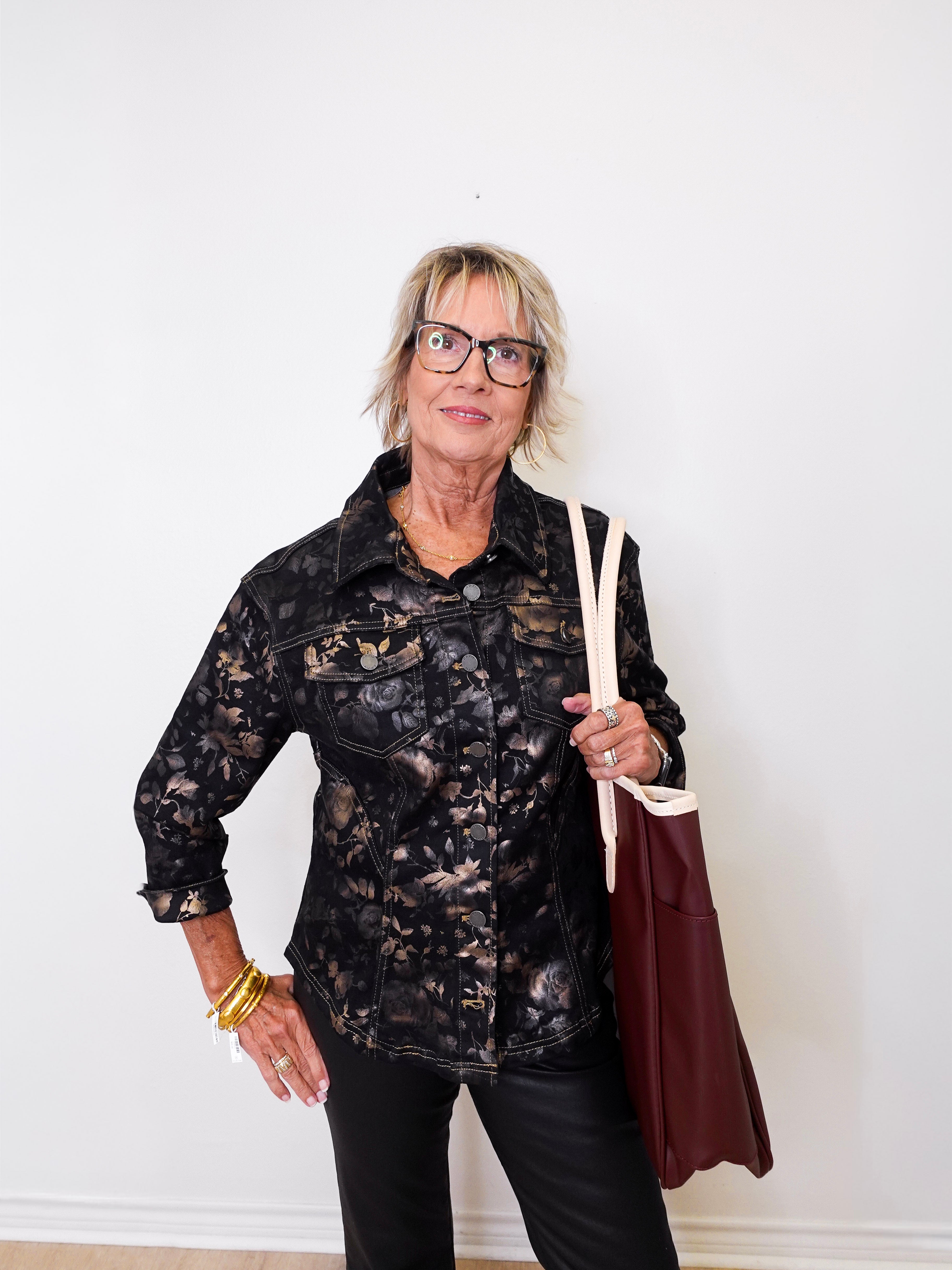 Woman wearing a black patterned shirt and holding a brown bag against a white wall