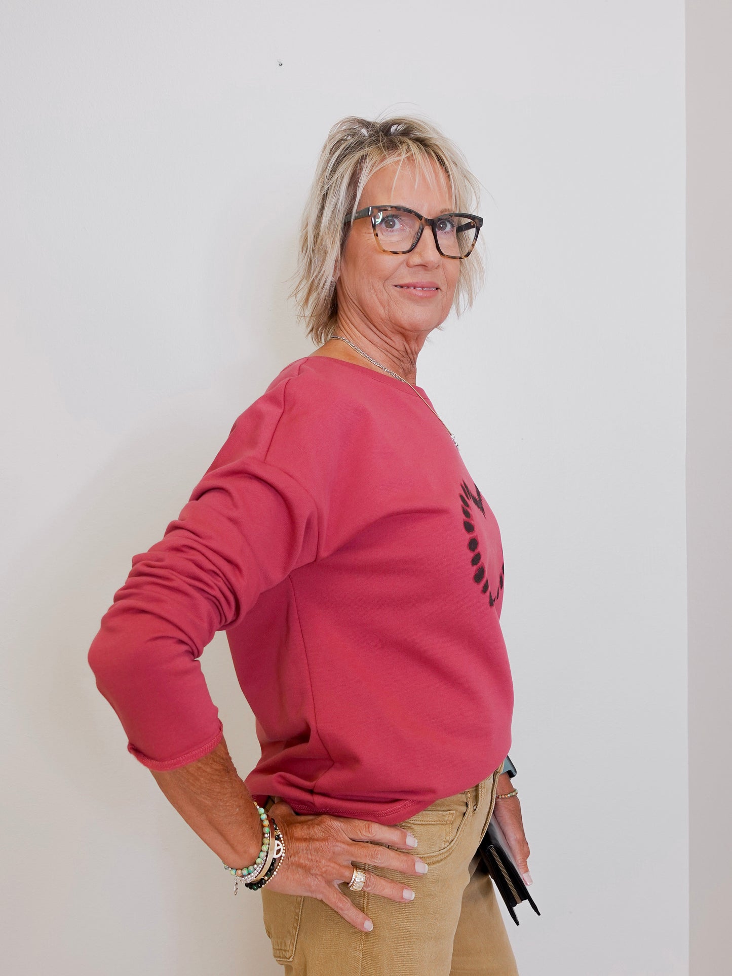 Woman wearing a pink sweatshirt with a logo, standing against a white wall.