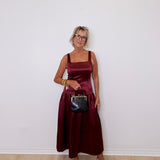 Woman in a burgundy dress holding a black handbag against a white wall.