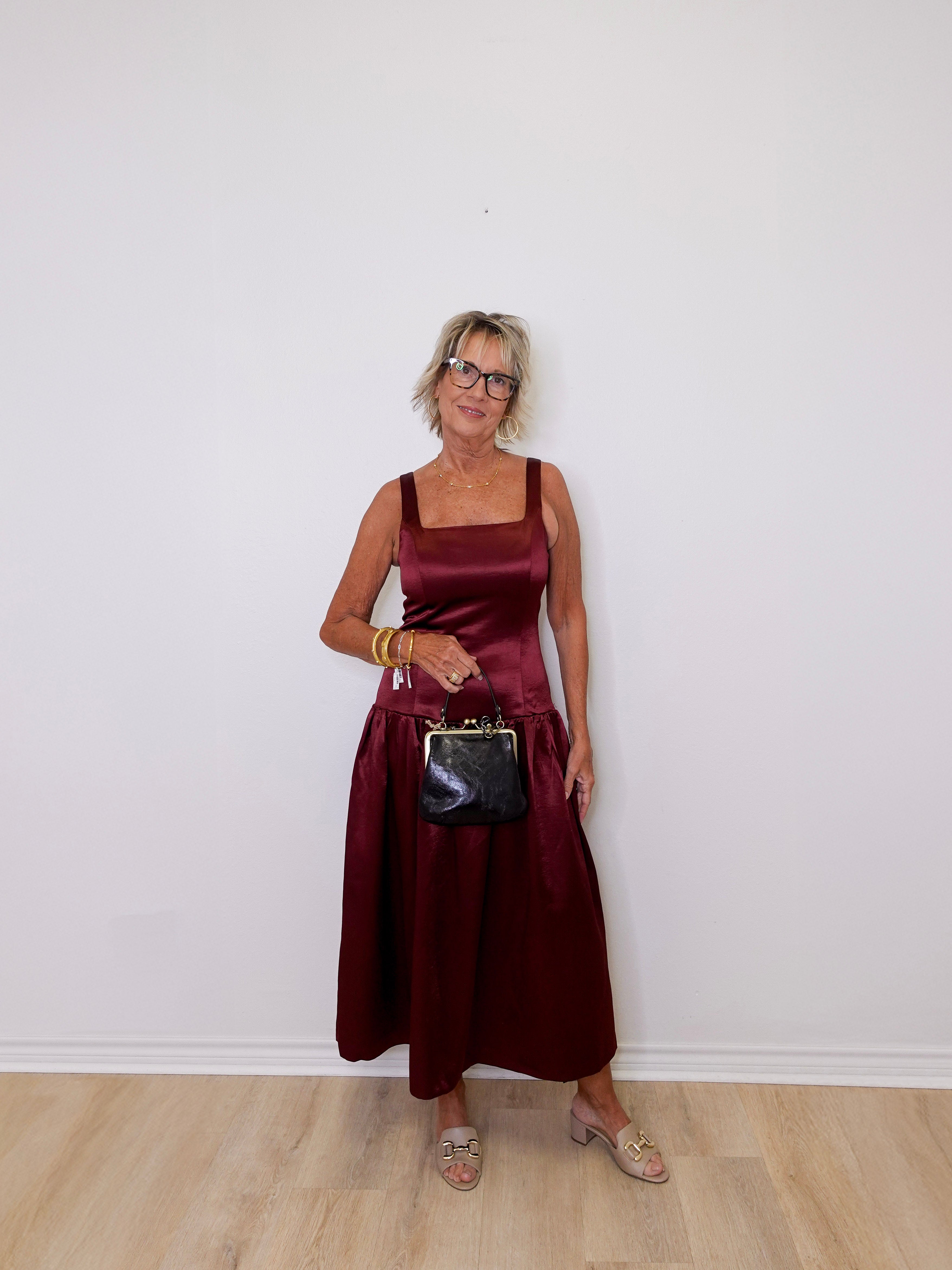 Woman in a burgundy dress holding a black handbag against a white wall.