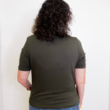 Person wearing an olive green t-shirt and blue jeans standing against a white wall.